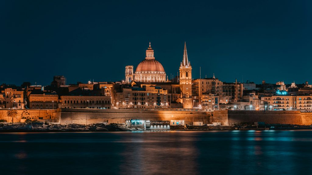 Valletta, Malta skyline at night across the harbor with illuminated historic buildings and domed church.