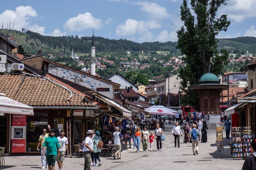 Bustling pedestrian street in Sarajevo’s Baščaršija with shops, cafes, and hills in the background on a sunny day