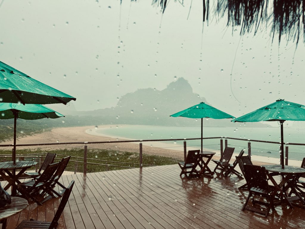 Rainy beach view from a seaside deck with green umbrellas, lounge chairs, and a quiet curved shoreline.