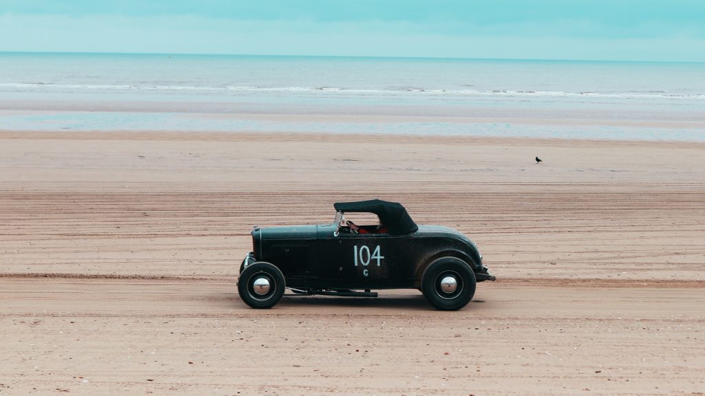Vintage black convertible driving along a wide, empty beach with the ocean in the background.