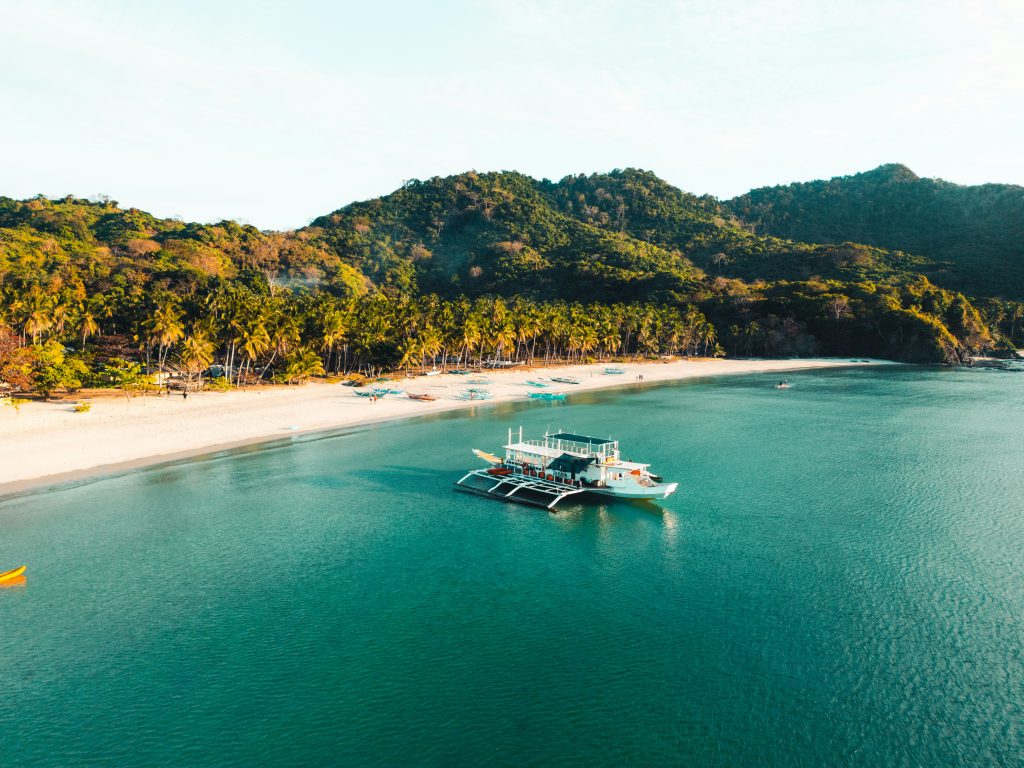 Aerial view of a white-sand beach with palm trees and an outrigger boat floating in clear turquoise water.