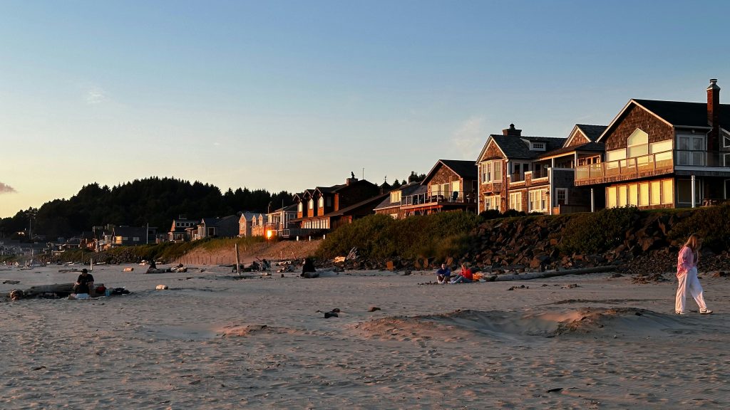 Beachfront houses glowing at sunset above a wide sandy beach with a few people walking and relaxing.